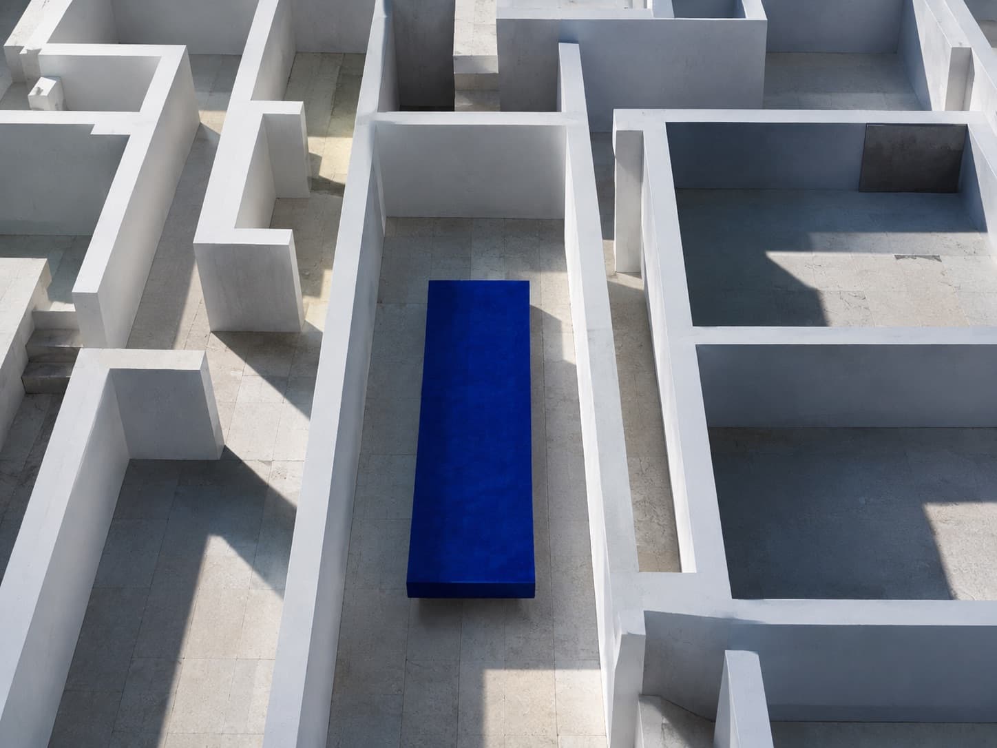 A white architectural maze seen from above, with a single cobalt bench at its centre