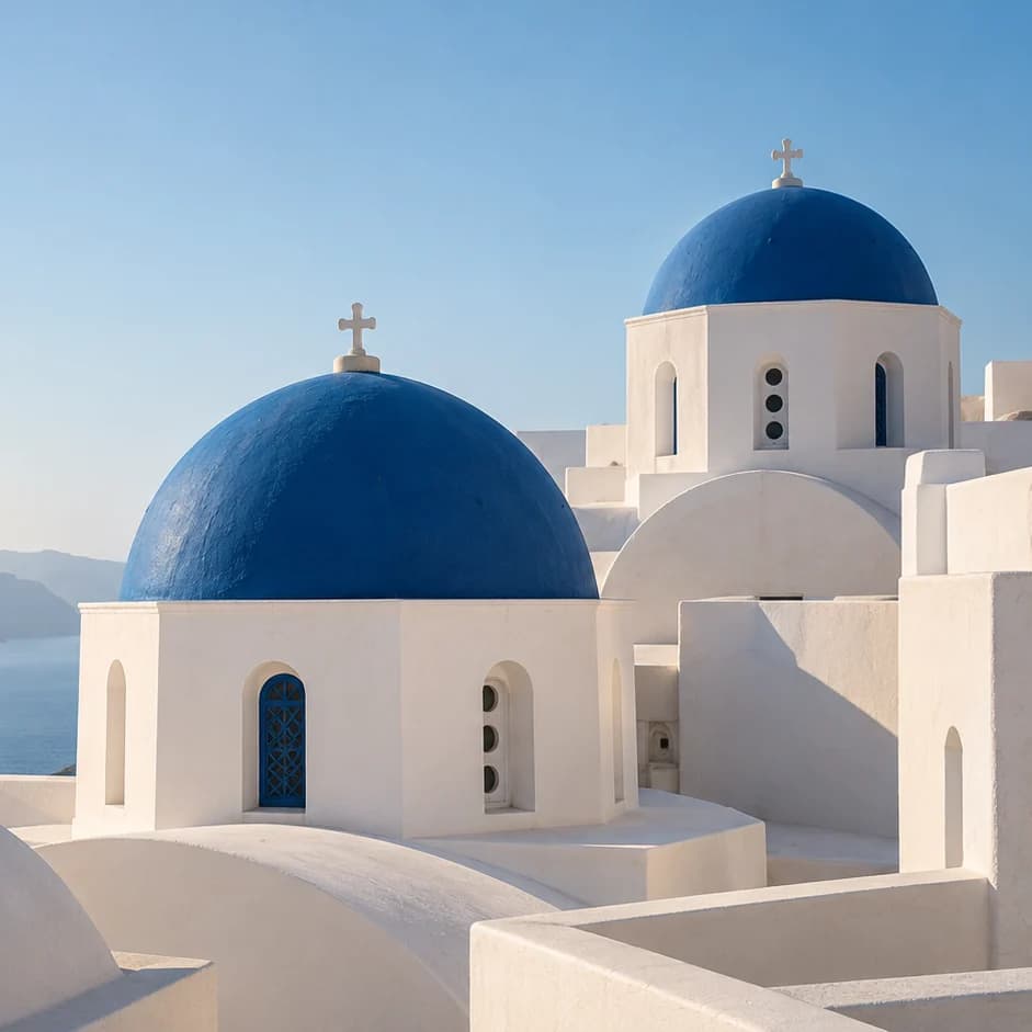 Two blue-domed white chapels at different heights above a pale sea, their rounded forms set among stepped whitewashed walls.