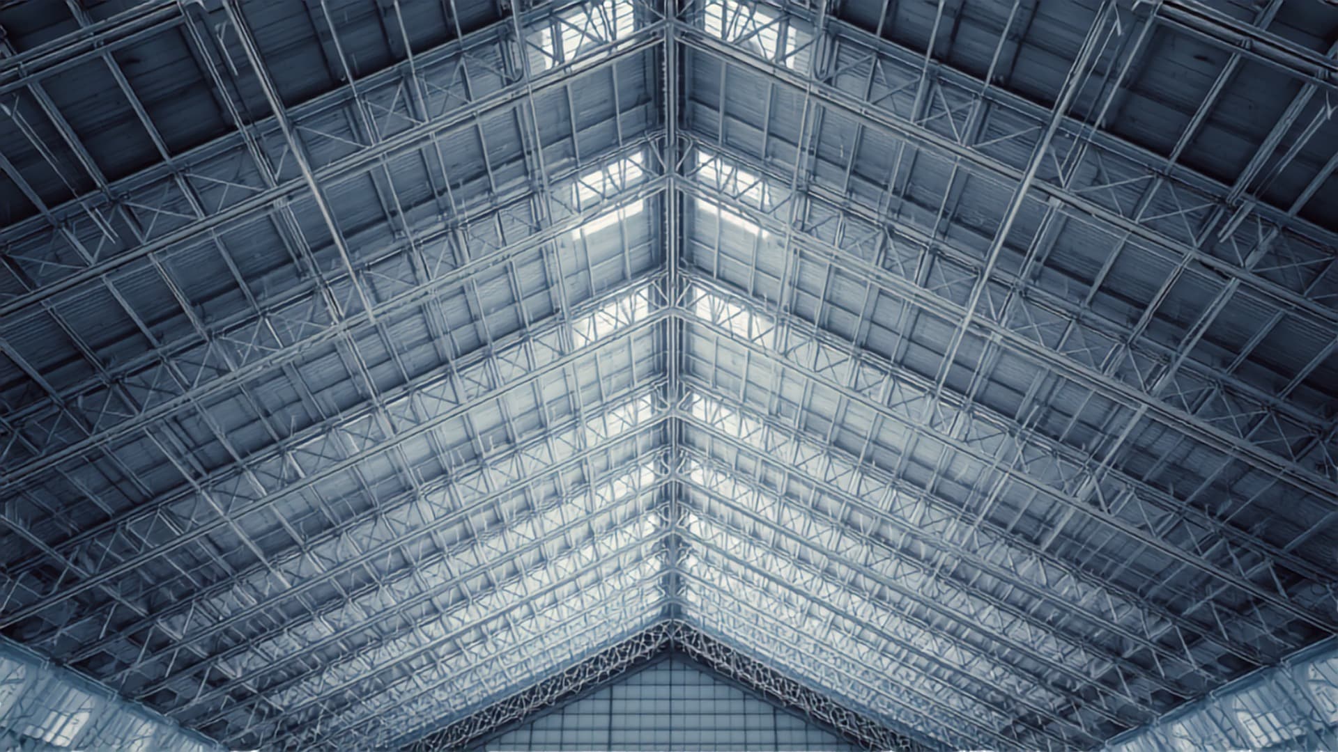 The triangulated steel roof structure of a large-span aircraft hangar, seen from below, daylight entering through the glazing.